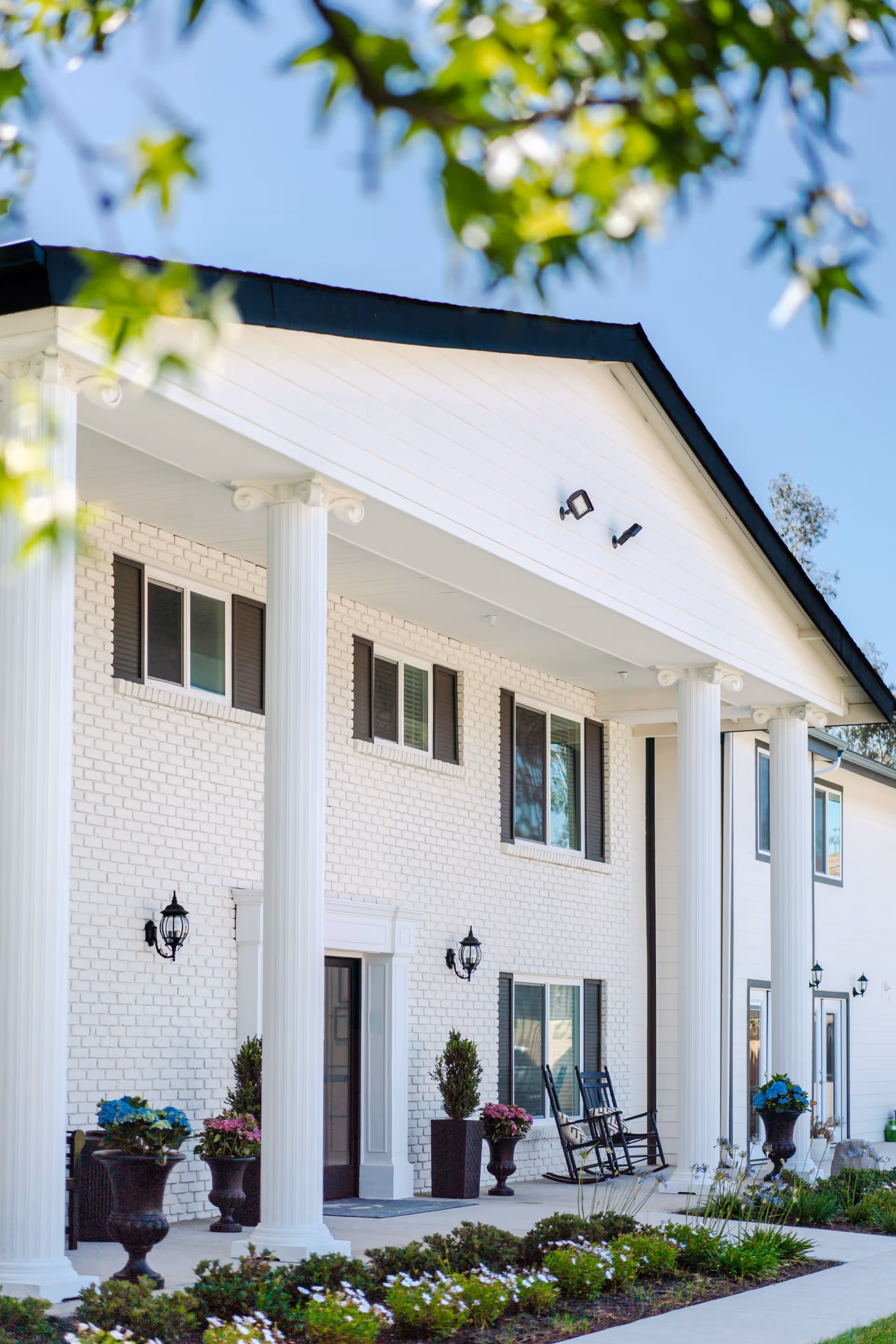 Front exterior view of a senior living facility with white brick walls, large white columns, black rocking chairs on the porch, potted plants, and a well-maintained garden under a clear blue sky.