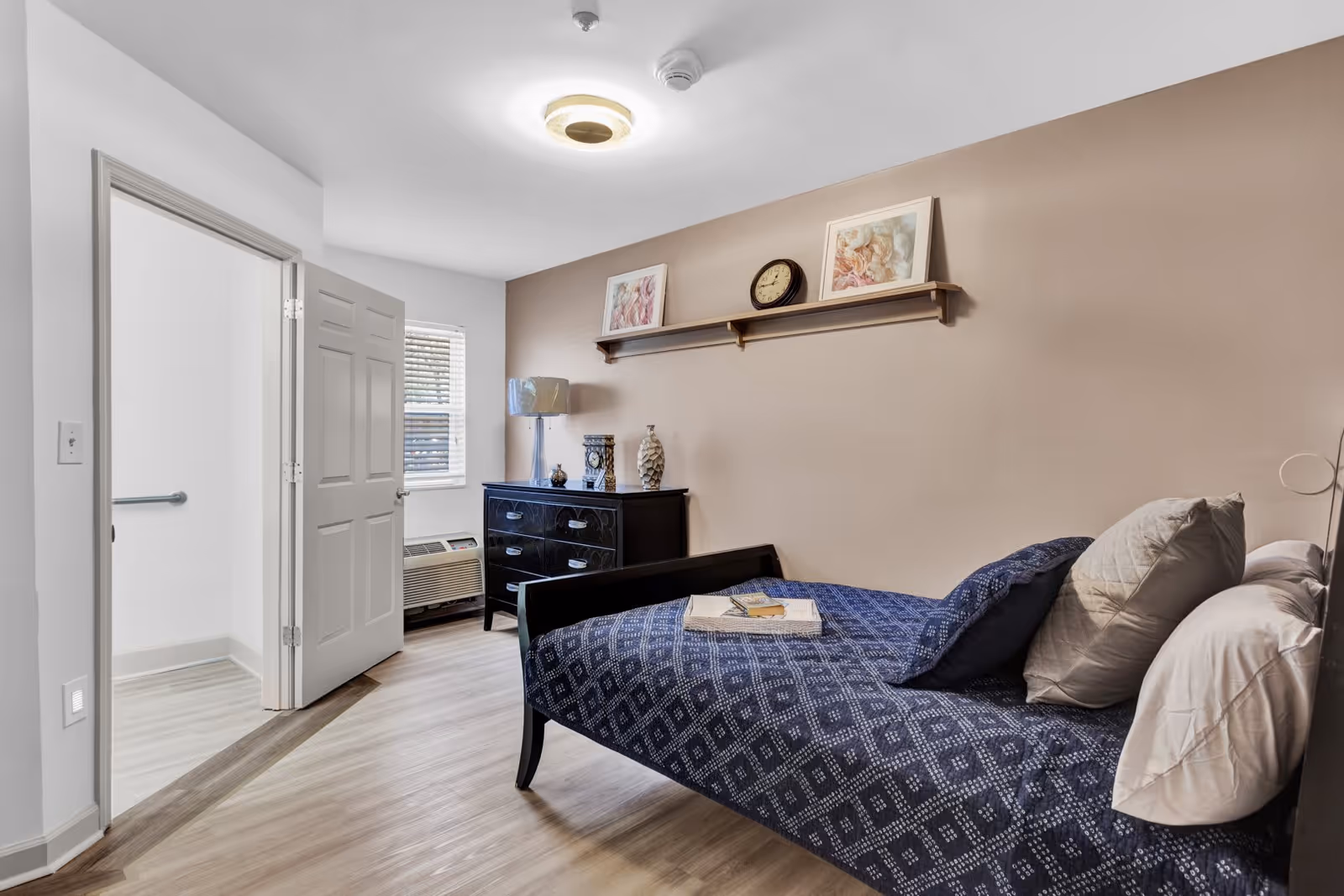 A neatly arranged bedroom in an assisted living facility featuring a single bed with blue patterned bedding and multiple pillows. A black dresser with decorative items and a lamp is positioned against a beige wall with a shelf above it holding two framed pictures and a clock. The room has light wood flooring and an open door leading to a bathroom with a grab bar visible.
