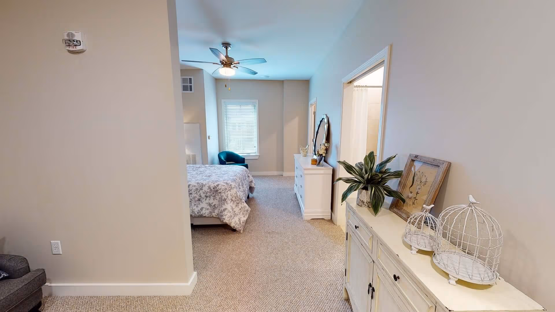 Interior view of a senior living facility bedroom with beige walls and carpeted floor. The room features a bed with floral bedding, a ceiling fan with light, a window with blinds, a blue armchair, a white dresser with a round mirror and decorative items, and a sideboard with a plant, framed artwork, and two decorative birdcages. A bathroom entrance is visible to the right.