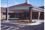 Entrance area of a senior living facility with a covered drop-off zone supported by brick columns, a landscaped flower bed in front, and a parking lot with a handicap parking space visible.