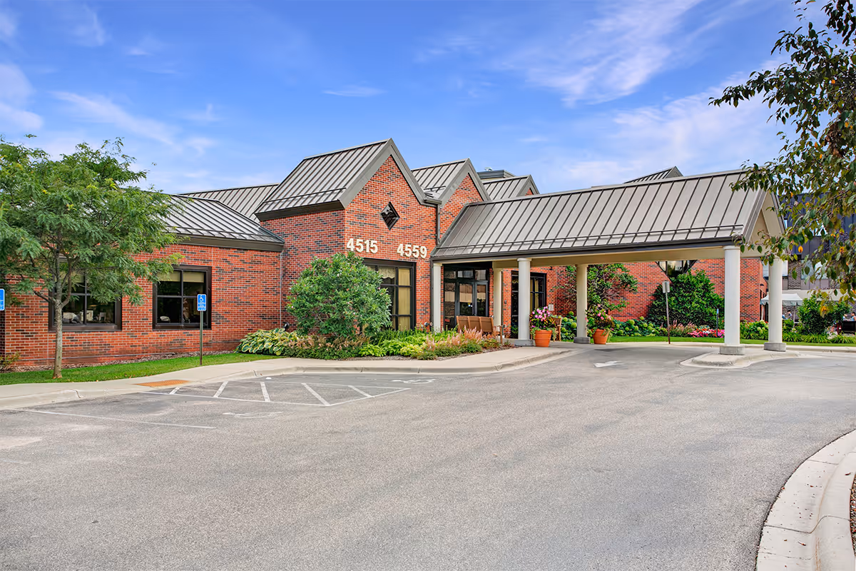 Exterior view of a brick building with metal roofing, featuring a covered entrance supported by white columns. The building has the numbers 4515 and 4559 displayed on the front. There are trees, shrubs, and potted plants around the entrance, and a clear blue sky above.
