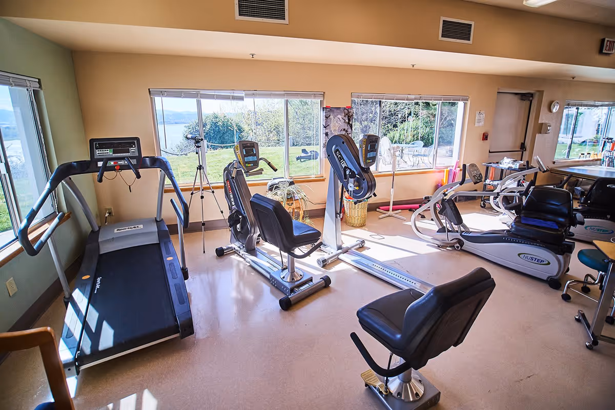 A bright exercise room with various fitness equipment including a treadmill, stationary bikes, and other workout machines. Large windows provide a view of greenery and a body of water outside. The room has beige walls and a light-colored floor.