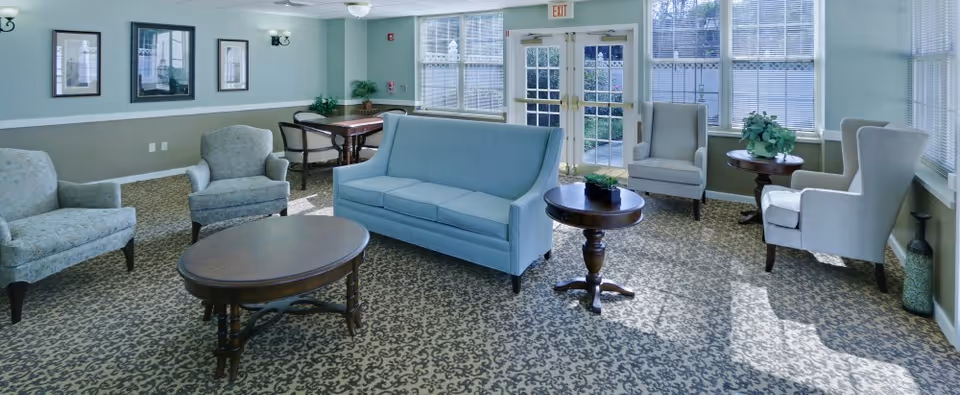 A bright and spacious living room area with patterned carpet, light blue sofa, several armchairs, wooden coffee tables, and large windows letting in natural light. There are framed pictures on the wall and a door leading outside.