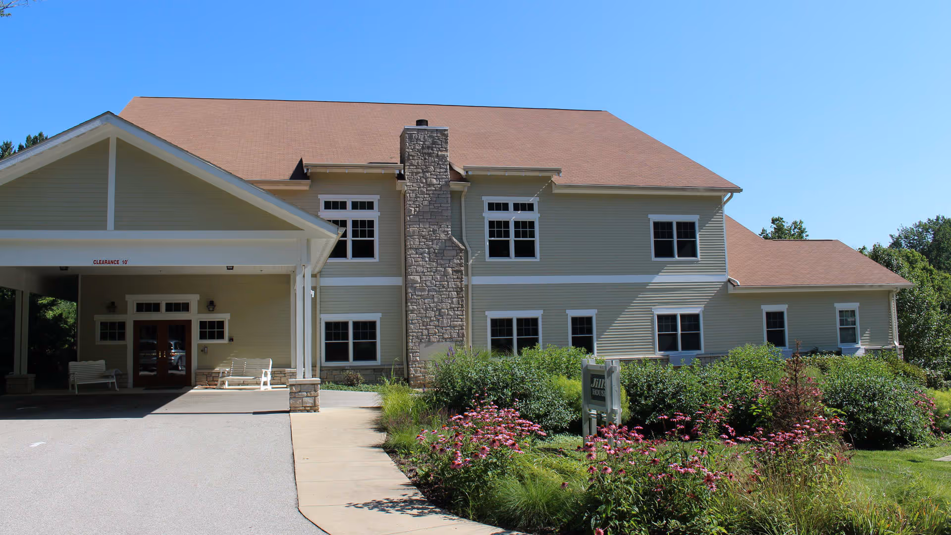 Exterior front view of a two-story beige building with a brown roof, stone chimney, and a covered entrance with benches. There is a garden with pink flowers and green shrubs in front of the building under a clear blue sky.