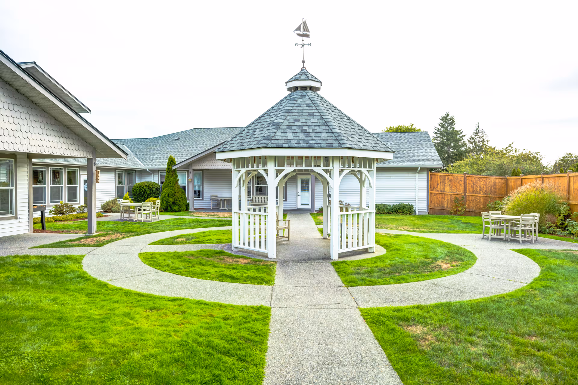 Outdoor courtyard area with a white gazebo in the center, surrounded by a circular concrete pathway and green grass. There are white buildings with gray roofs in the background, along with outdoor tables and chairs on the grass near the buildings. A wooden fence and some trees are visible in the background.