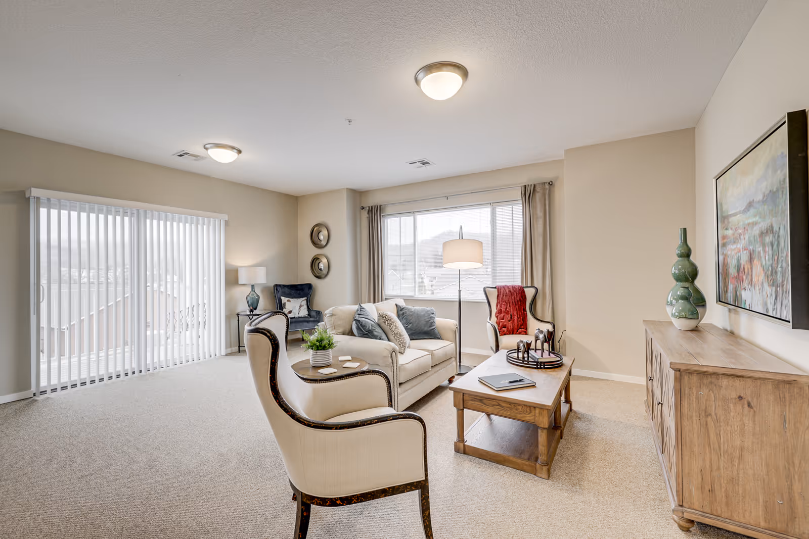 A bright and spacious living room with beige walls and carpet. The room features a cream-colored sofa with blue and patterned pillows, two armchairs, a wooden coffee table with decorative items, a wooden sideboard with green vases, a large window with curtains, and sliding glass doors with vertical blinds. There are two ceiling lights and a floor lamp near the window.