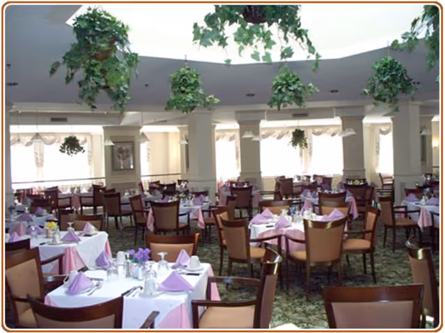 Spacious dining room with multiple tables set with pink tablecloths and folded napkins, surrounded by chairs and hanging plants.