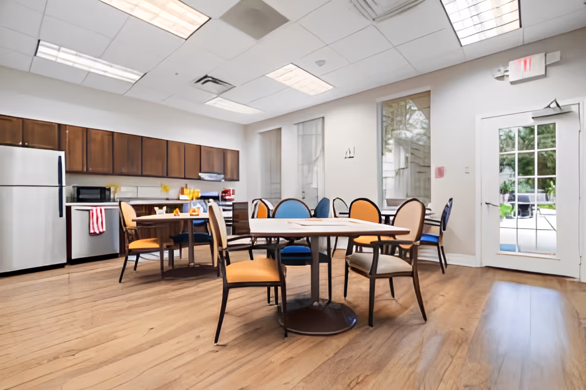 Bright communal dining area with round tables and colorful chairs, a kitchenette with cabinets and appliances, and a door to the outside.