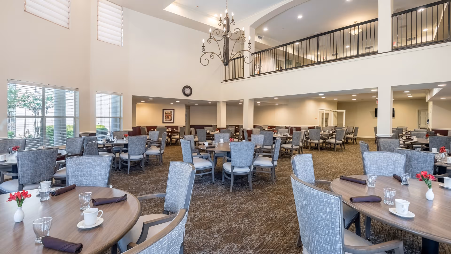 Spacious dining room with multiple round tables set with cups, glasses, and napkins. The room features high ceilings, large windows allowing natural light, and a decorative chandelier hanging from the ceiling. There is a second-floor balcony overlooking the dining area.