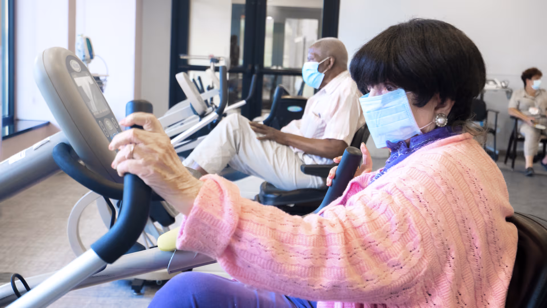 Two elderly individuals wearing face masks exercising on stationary bikes in a fitness room with large windows and other people seated in the background.