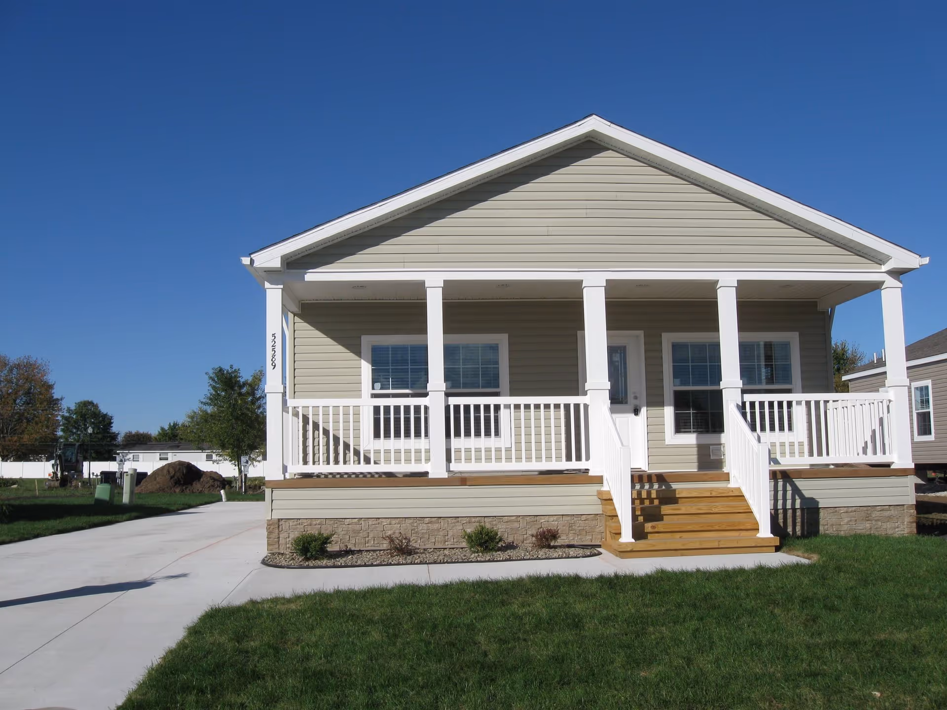 Front exterior view of a single-story house with a covered porch, white railings, and steps leading up to the front door. The house has beige siding and a stone foundation, with a clear blue sky in the background and a green lawn in the foreground.