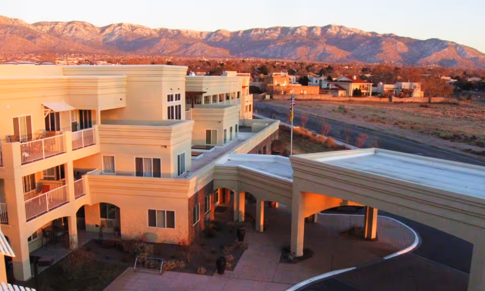 Exterior view of a multi-story beige senior living building with balconies, a covered entrance drive, and mountains in the background at sunset.
