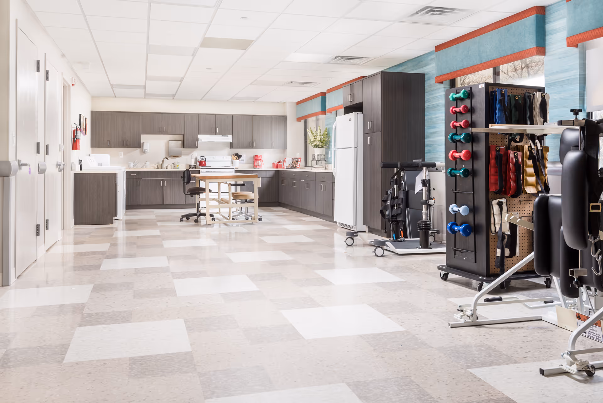 A bright rehabilitation room with a kitchenette along the back wall and various therapy equipment and weights in the foreground.
