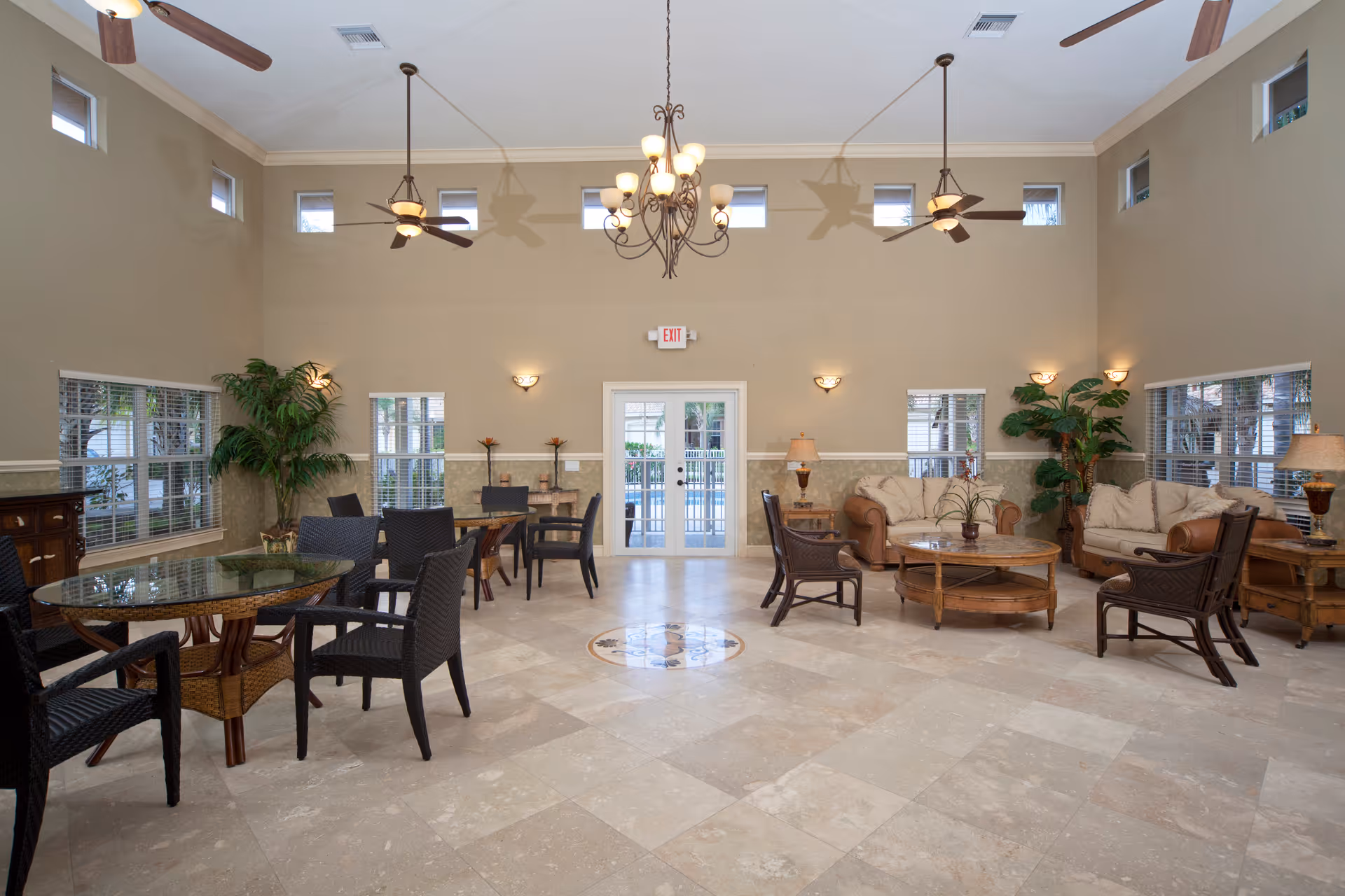Spacious common area with high ceilings, ceiling fans, and multiple windows. The room features several seating arrangements including glass-top tables with black chairs and a lounge area with beige sofas, armchairs, wooden coffee tables, and potted plants. There is a double glass door at the far end of the room.