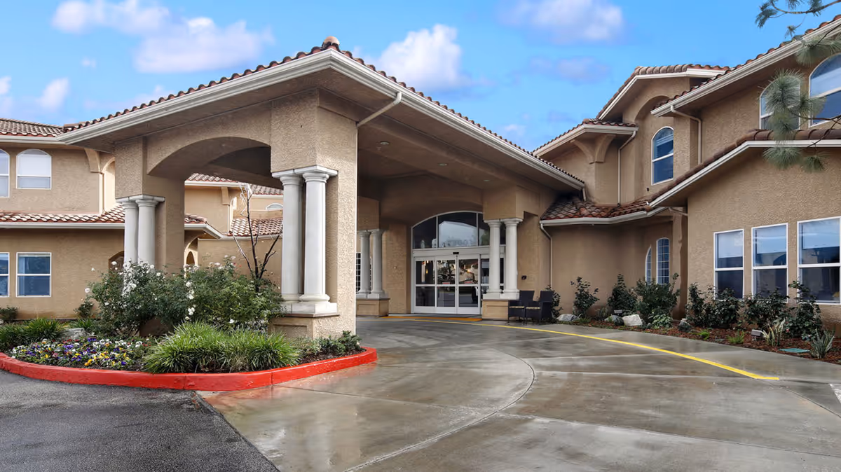 Exterior view of Ivy Park at Simi Valley senior living facility entrance with a covered driveway, beige stucco walls, red-tiled roof, white columns, and landscaped flower beds.