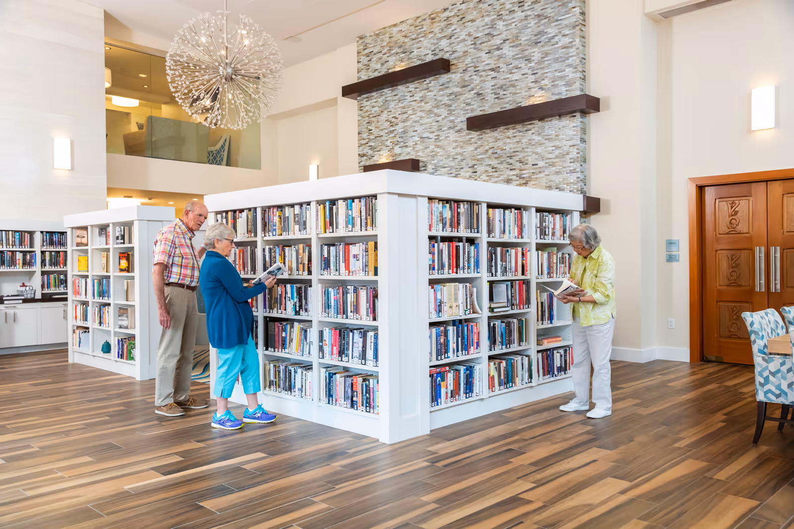 Three older adults browse books on white shelves in a bright, modern library area of a senior living facility.