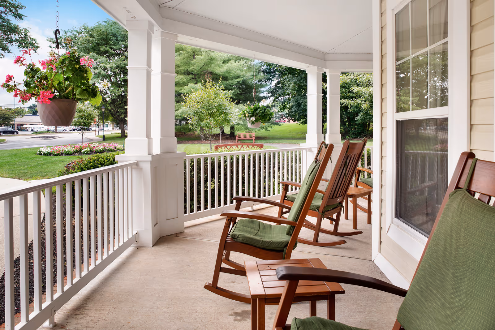 Covered porch with wooden rocking chairs featuring green cushions, small wooden tables, white railing, hanging flower pot with pink flowers, and a view of green trees and a bench in the background.