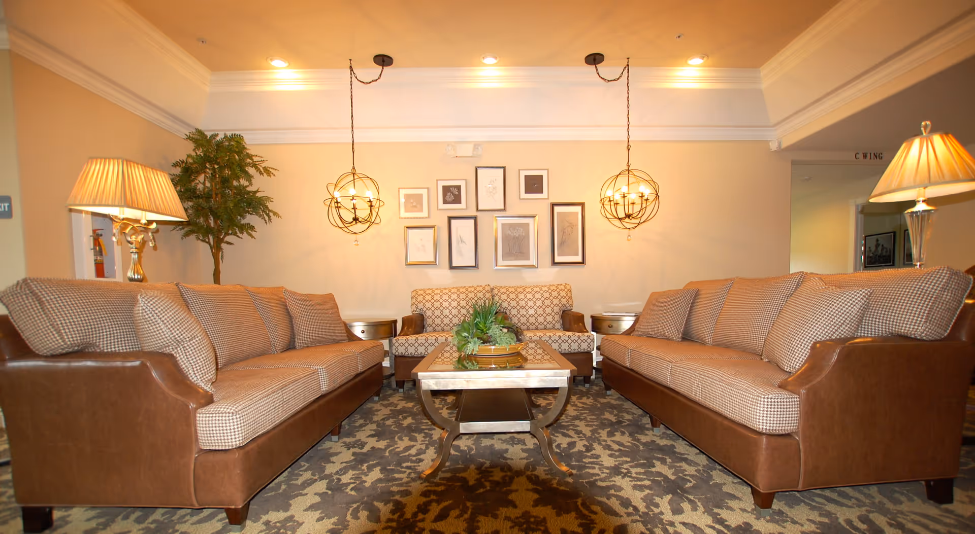 Communal living room with two upholstered sofas facing a central coffee table, pendant lights, framed art, and lamps.