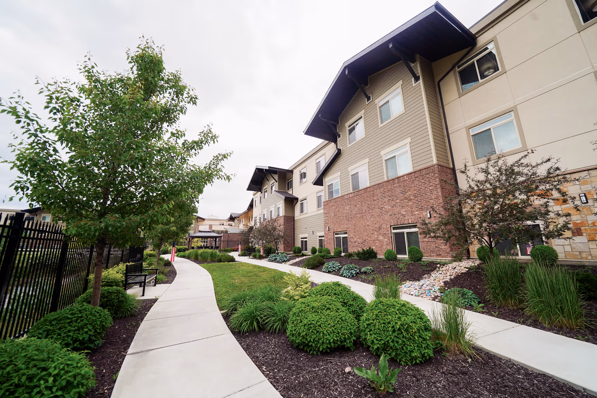 A paved walkway curves through a landscaped garden area with green bushes, trees, and mulch alongside a multi-story senior living facility building with beige and brick exterior walls under a cloudy sky.