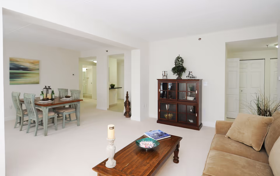A bright and spacious living and dining area in a senior living facility. The living room features a beige sofa with matching cushions, a wooden coffee table with a candle and decorative bowl, and a wooden cabinet with glass doors displaying various items. The dining area has a wooden table with six chairs, set with plates and wine bottles. The walls are painted white, and there is a large abstract painting hanging above the dining table. The space is carpeted in light beige, and there are doorways leading to other rooms.