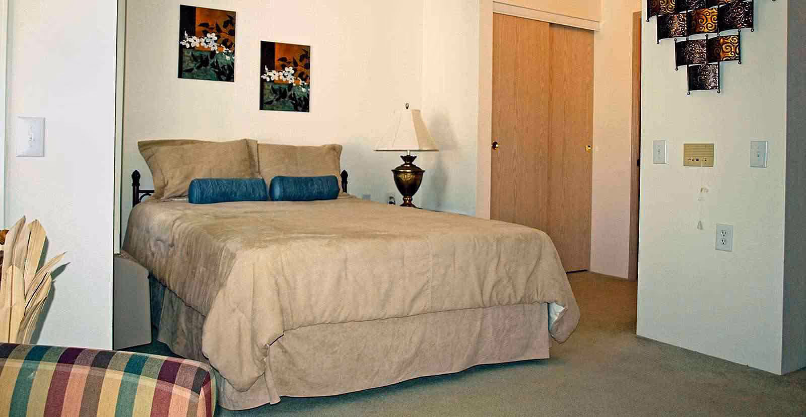 A bedroom with a beige bedspread and two blue cylindrical pillows on the bed. There is a bedside table with a lamp, two floral paintings on the wall above the bed, a closet with sliding wooden doors, and a striped armchair partially visible in the foreground.