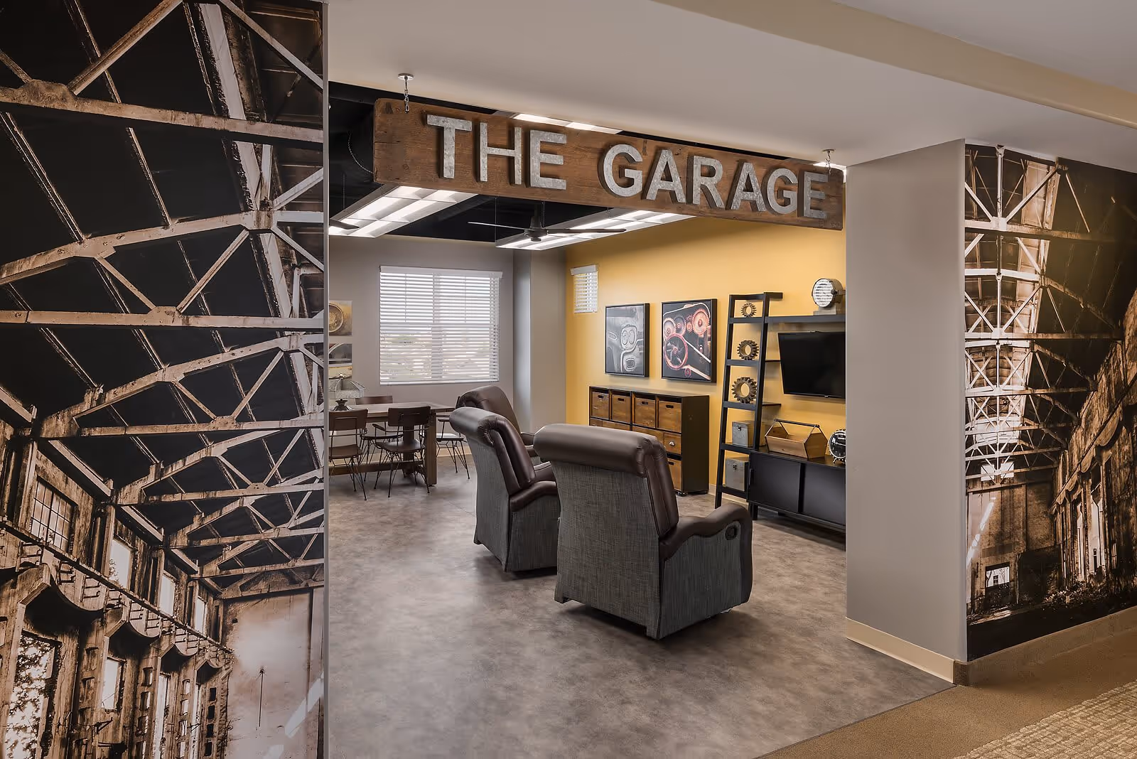 Interior common lounge labeled "The Garage" with two recliners facing a TV, shelving, and a table near a window.