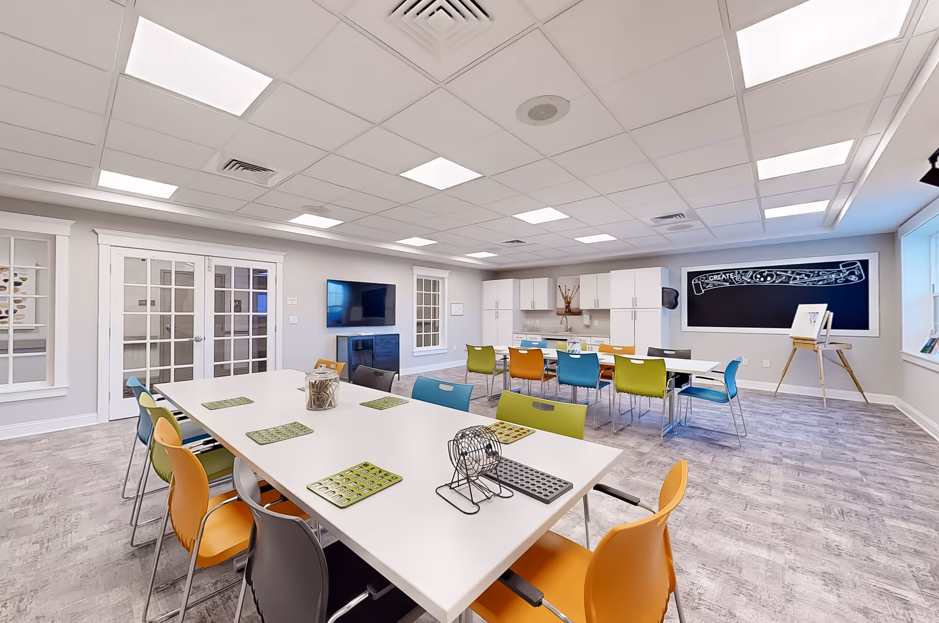 A bright, spacious activity room with two white tables surrounded by colorful chairs in orange, green, blue, and gray. On the tables are bingo cards and a bingo cage. The room has a light gray floor, white walls, and a ceiling with recessed lighting. There is a large blackboard with the word 'CREATE' and artistic designs on the far wall, an easel with a canvas, a flat-screen TV mounted on the wall, and white cabinets with a sink area in the back.