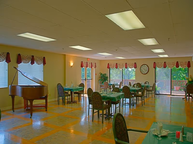 A spacious dining room with several tables covered in green tablecloths and set with plates, glasses, and napkins. The room features large windows and sliding glass doors with red and white valances, allowing natural light to fill the space. A grand piano is positioned near the windows on the left side. The floor has a patterned design, and a large clock is mounted on the far wall.