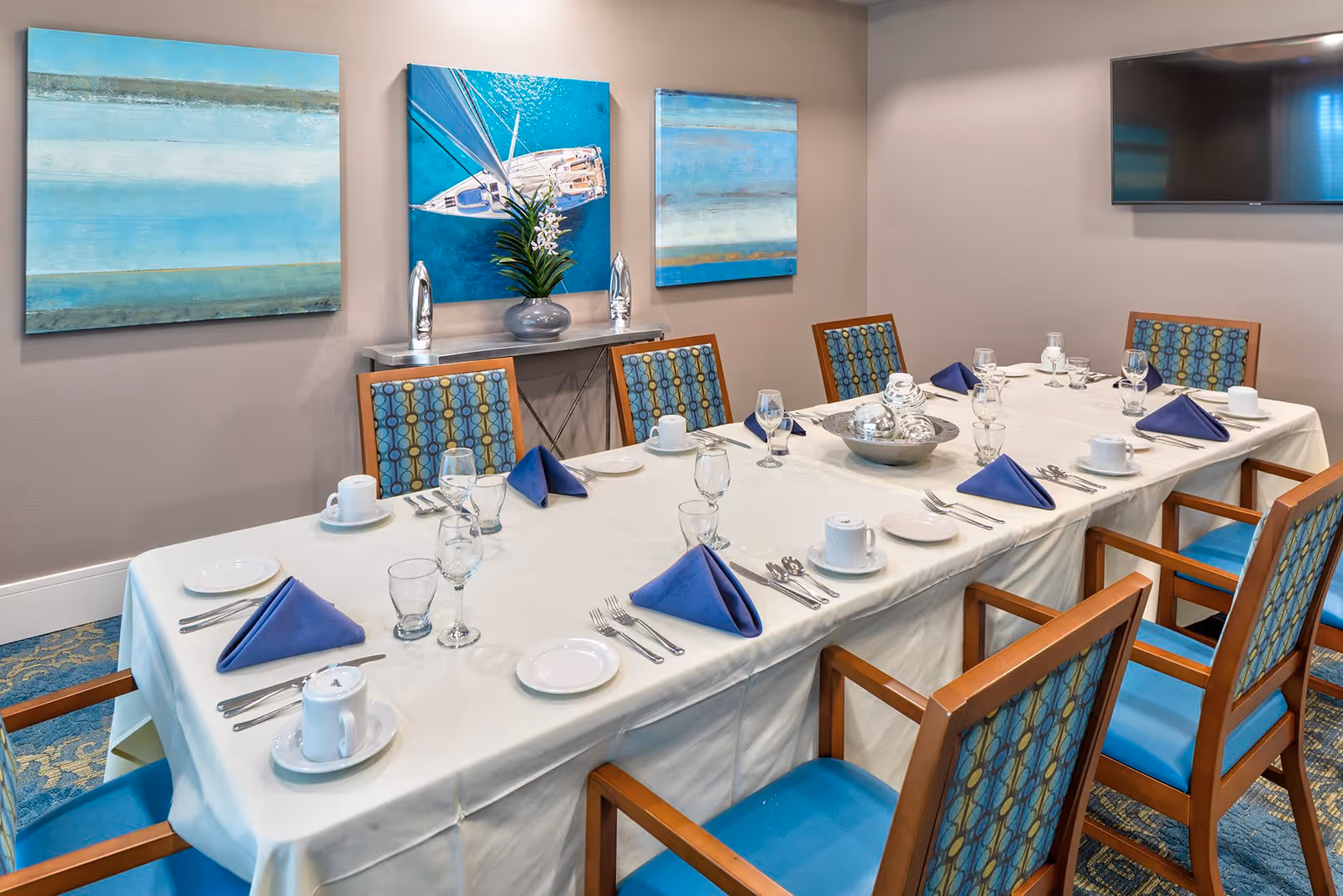 A dining room with a long rectangular table set for eight people. The table is covered with a white tablecloth and has blue folded napkins, white plates, cups, glasses, and silverware arranged neatly. The chairs have wooden frames with blue cushioned seats and patterned backs. On the wall behind the table are three blue-themed paintings and a small console table with decorative items and a plant. A flat-screen TV is mounted on the adjacent wall.