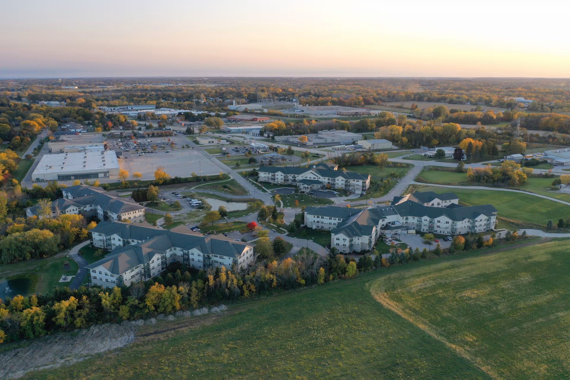 Aerial view of the Parkview Gardens campus showing multiple connected senior living buildings surrounded by trees, roads, and open fields at sunset.