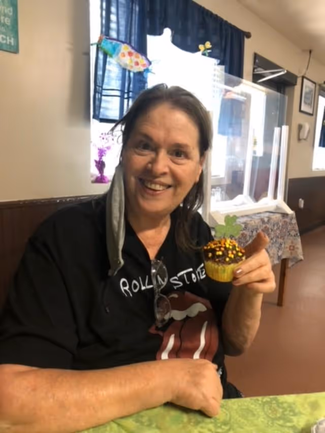 A smiling woman sitting at a table indoors, holding a decorated cupcake with autumn-themed sprinkles and a green leaf topper. She is wearing a black Rolling Stones t-shirt and has a gray face mask hanging from one ear. Behind her, there is a window with blue curtains and some colorful decorations.