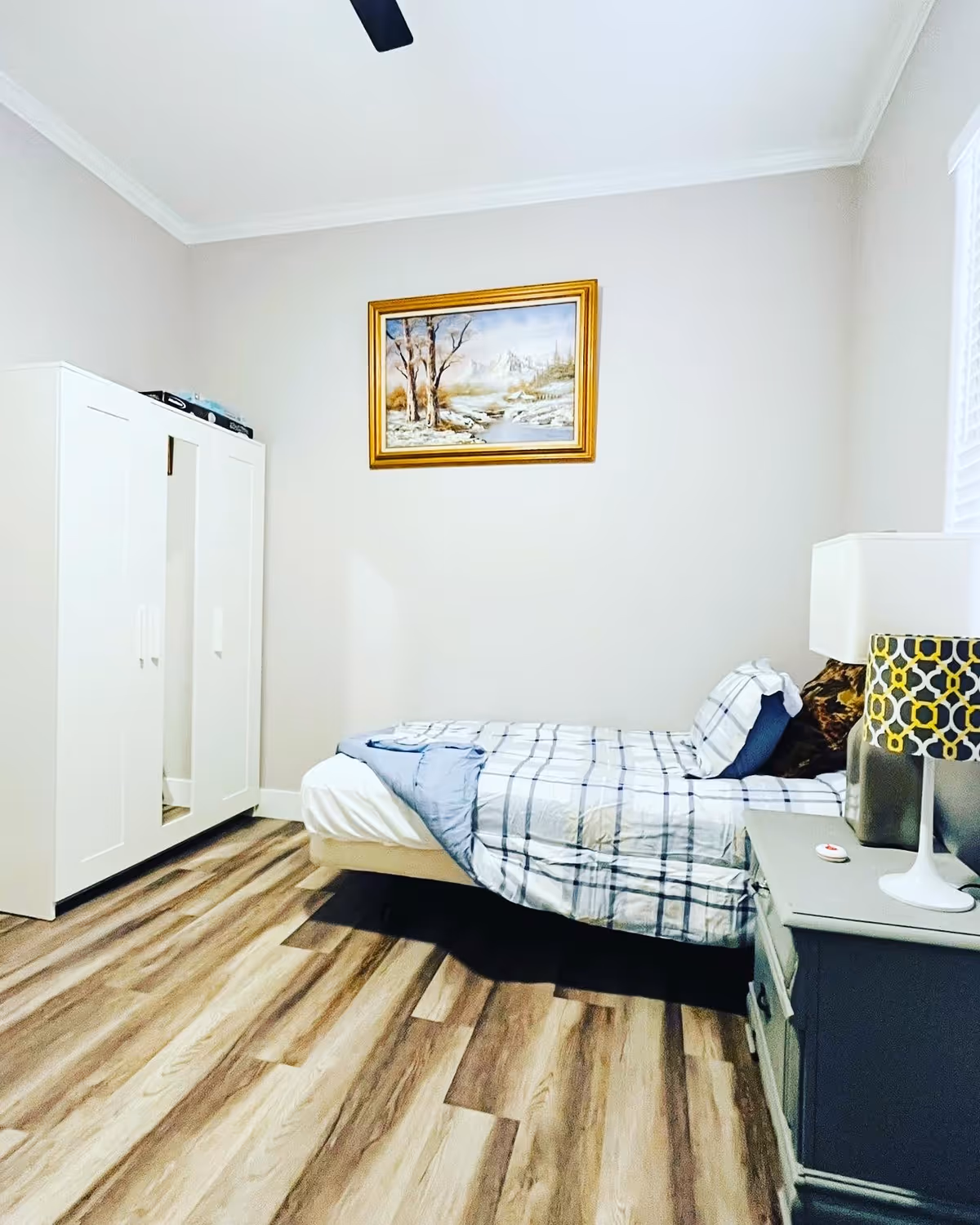 A neatly arranged bedroom with a single bed covered in a blue and white checkered blanket and pillows. Next to the bed is a gray nightstand with a white lamp featuring a patterned lampshade. A white wardrobe with a mirror on one door stands against the wall. Above the bed hangs a framed painting of a landscape with trees and mountains. The room has wood-patterned flooring and light-colored walls.