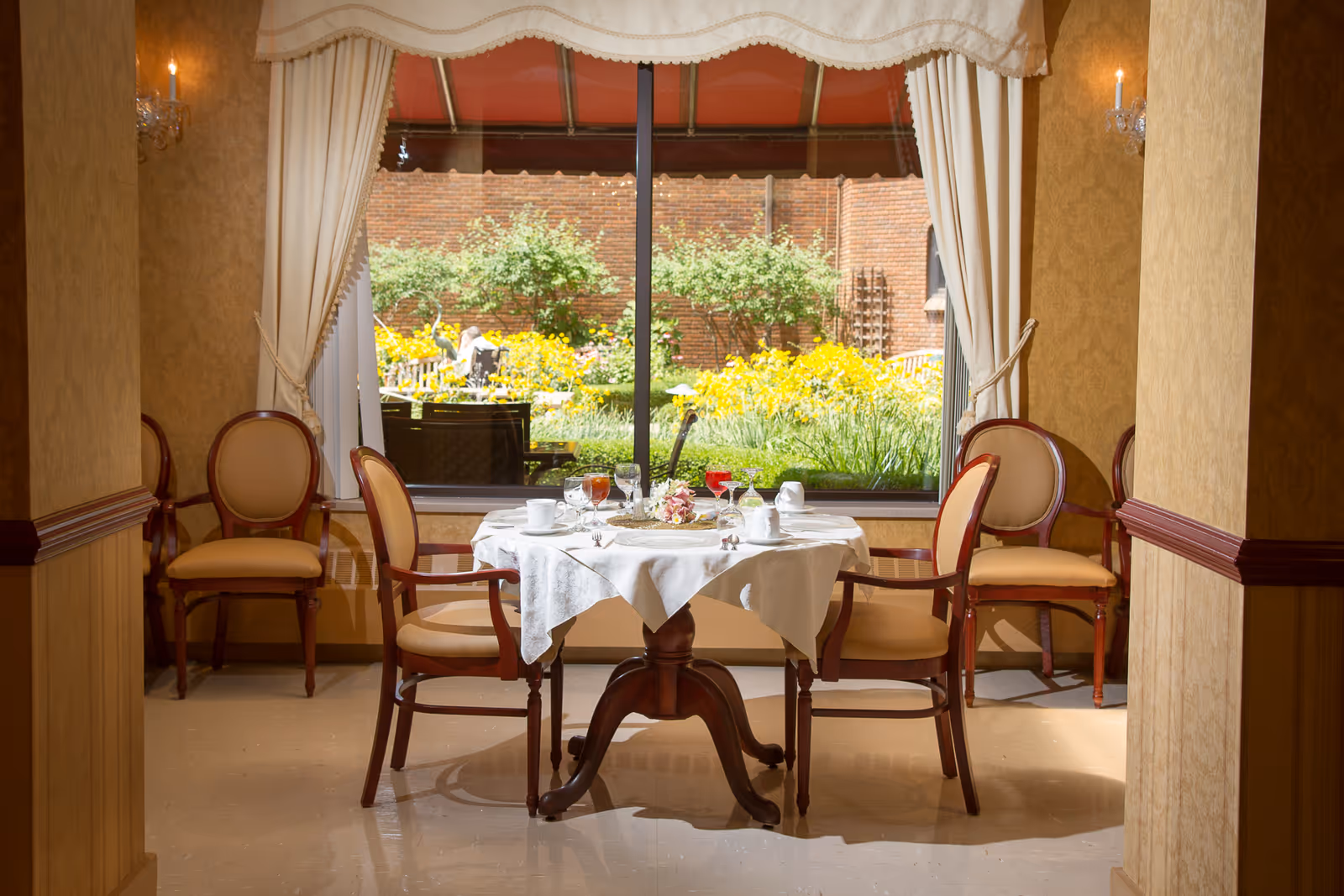 A small dining area with a round table covered with a white tablecloth set for four people. The table has cups, glasses with beverages, and a floral centerpiece. Four wooden chairs with cushioned seats surround the table. Behind the table is a large window with cream-colored curtains, showing a garden with yellow flowers and green shrubs outside. The room has warm beige walls and soft lighting from wall sconces.