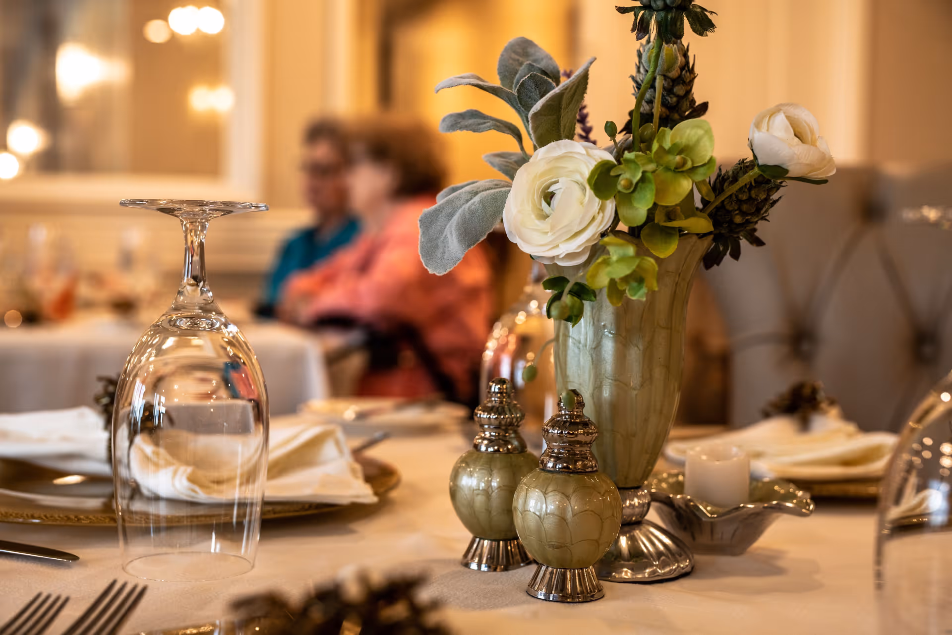 Close-up of a dining table set with an upside-down wine glass, folded napkins on plates, salt and pepper shakers, a small candle, and a vase with white and green flowers. In the background, two elderly people are sitting and conversing in a softly lit room.