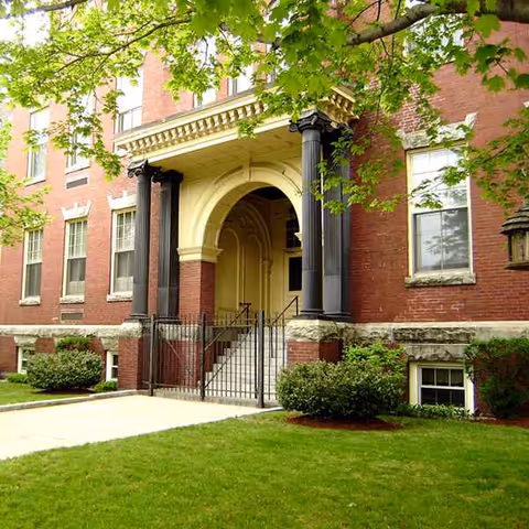Brick assisted living building entrance with arches and columns, green lawn and trees.