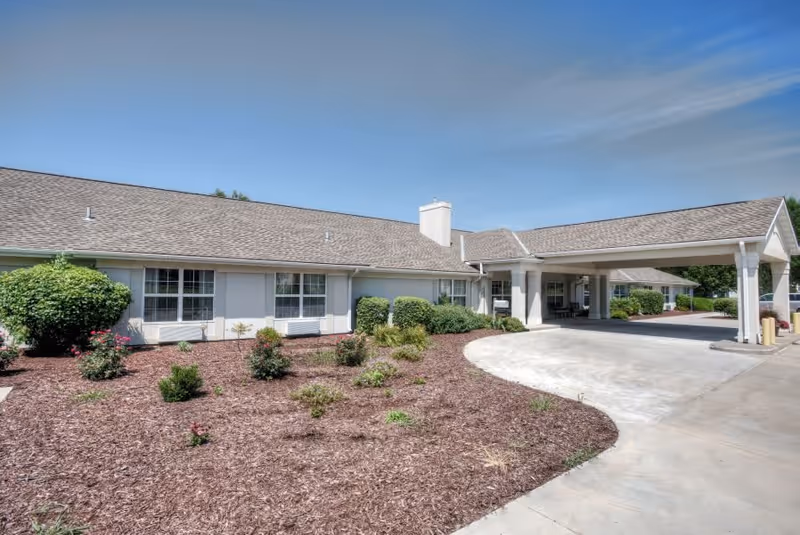 Exterior view of a single-story senior living facility building with a covered entrance driveway, landscaped with small bushes and mulch under a clear blue sky.