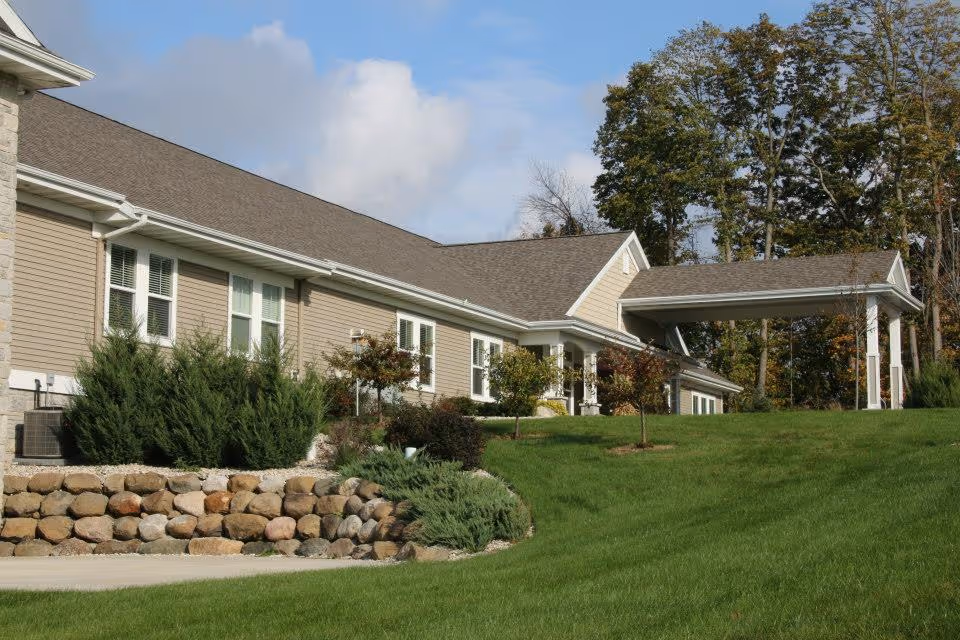 Exterior view of a single-story assisted living facility building with beige siding, multiple windows, a covered entrance, and a well-maintained lawn with shrubs and small trees.
