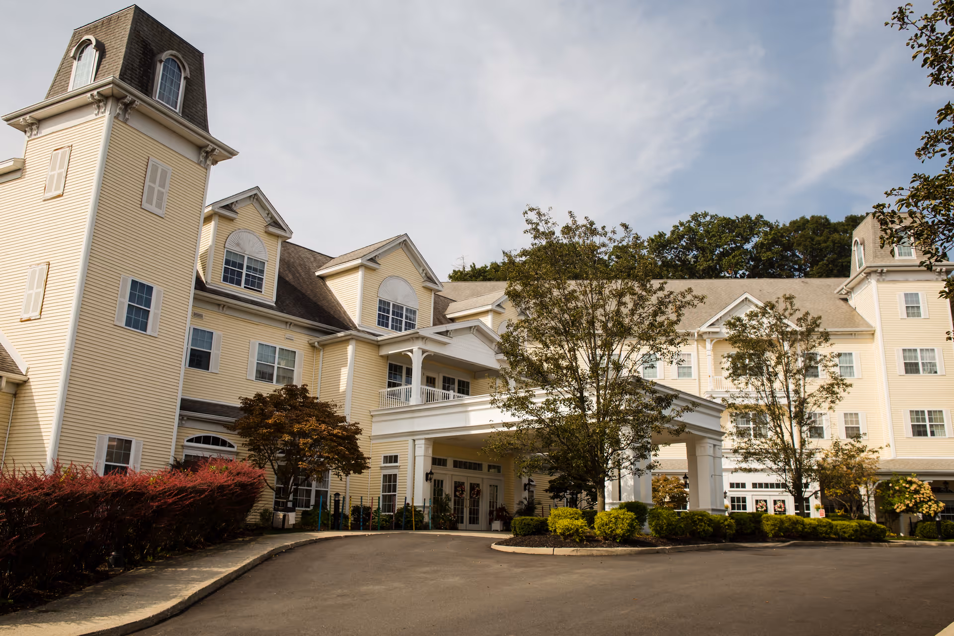 Exterior view of a large, multi-story senior living facility building with beige siding, multiple windows, and a covered entrance. There are trees and shrubs around the driveway leading to the entrance under a partly cloudy sky.