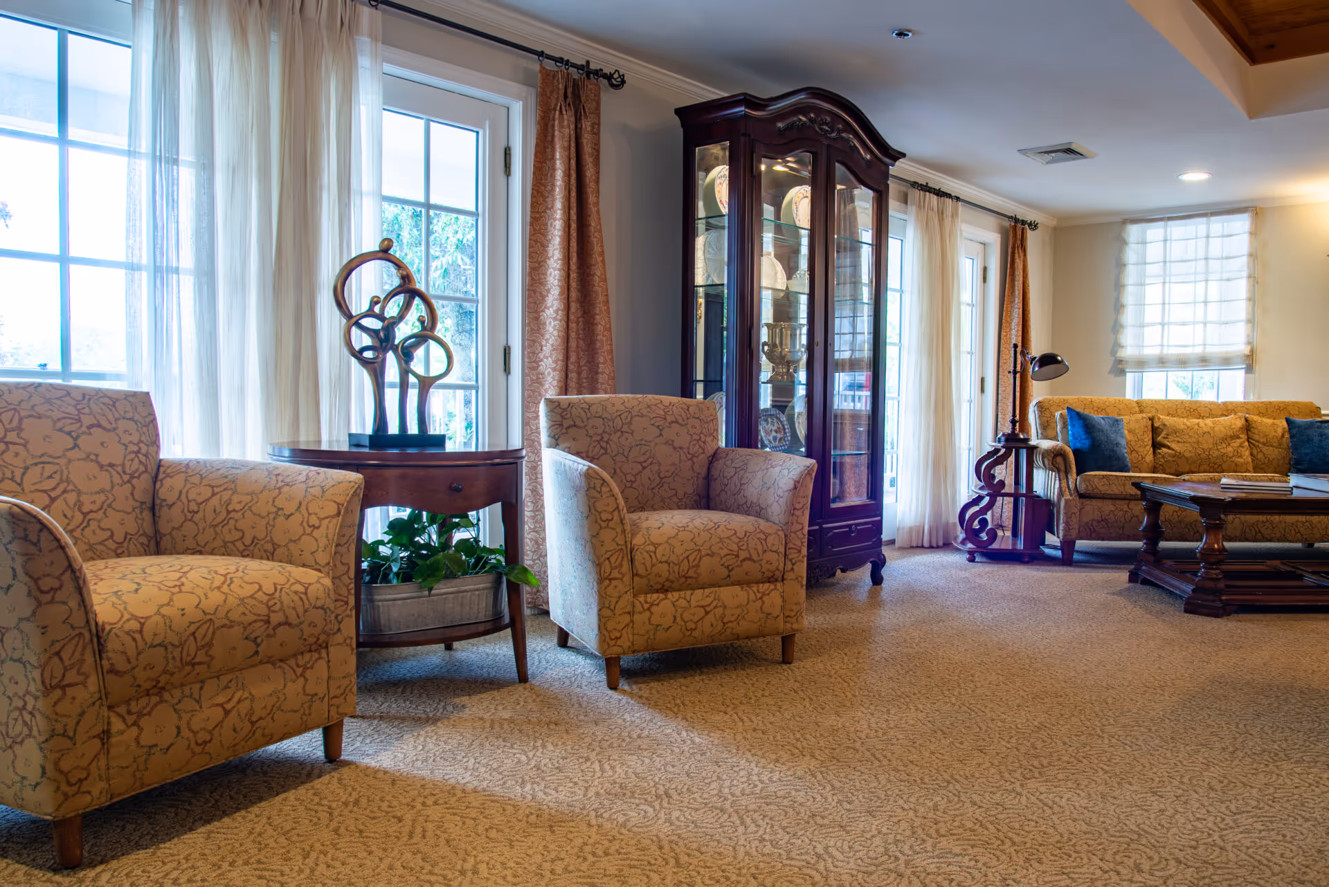 A cozy living room with two patterned armchairs, a wooden side table with a decorative sculpture, a glass-fronted cabinet displaying plates and trophies, a sofa with blue cushions, a wooden coffee table, and large windows with sheer and opaque curtains letting in natural light.