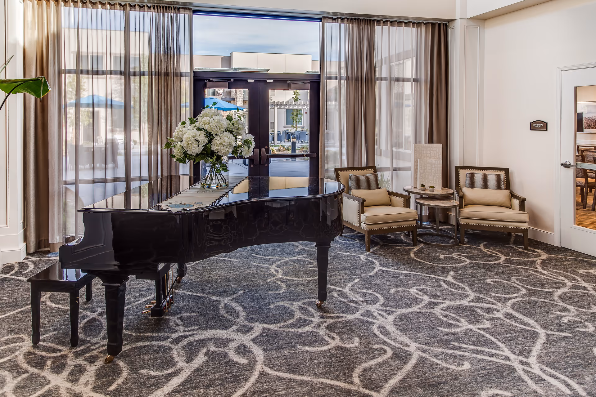A bright and elegant interior room featuring a black grand piano with a floral arrangement on top, two beige armchairs with cushions, a small round side table, large windows with sheer curtains, and a door leading to an outdoor area with umbrellas.