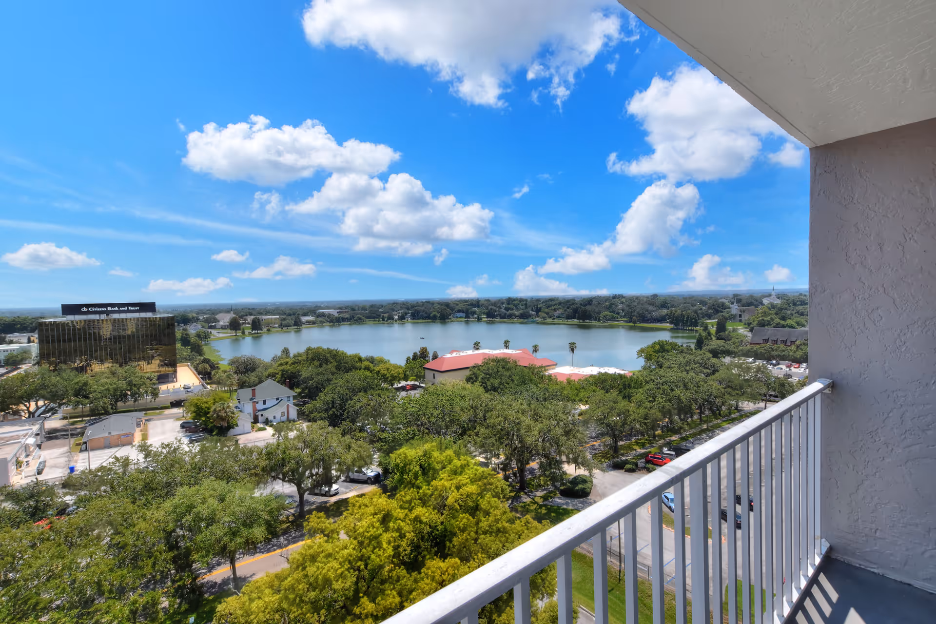 View from a balcony showing a railing overlooking trees, a lake, and nearby buildings under a blue sky with clouds.