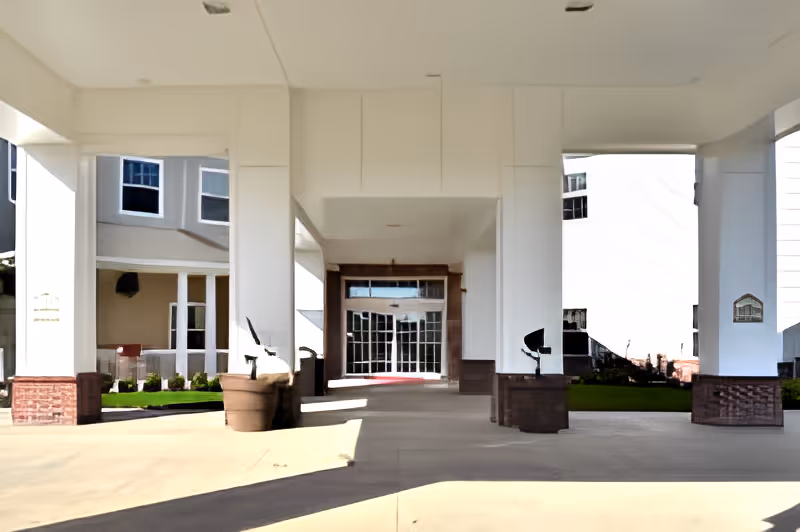 Covered entrance area of a building with white pillars and a glass door at the end, surrounded by parts of the building with windows and some greenery on either side.