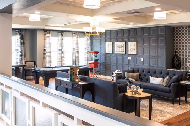 A spacious and well-lit common area with a pool table, dark blue sofas, a small round table with decorative items, and a popcorn machine. The room features large windows with patterned curtains, a dark paneled accent wall with framed artwork, and modern ceiling lights.