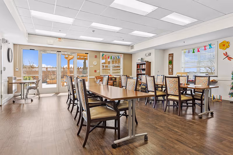 A bright and spacious dining room with multiple wooden tables and chairs arranged neatly. Large windows and glass doors allow natural light to fill the room, showing an outdoor patio area. The walls are decorated with colorful paper cutouts and a wooden sign that reads 'HOME'. The floor is wooden, and there is a bookshelf and some small decorations in the corner.