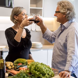 An older woman and man smiling and toasting with glasses of red wine in a kitchen setting, with fresh vegetables and a bottle of wine on the counter in front of them.