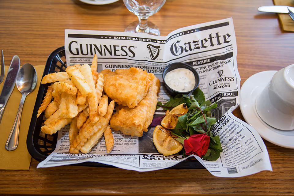 A serving of fish and crinkle-cut fries on a tray lined with a newspaper-style paper featuring the Guinness Gazette. The meal includes two pieces of fried fish, a small cup of tartar sauce, grilled lemon wedges, and a garnish of greens with a red flower. The tray is on a wooden table with a fork, spoon, knife, and a white cup and saucer nearby.