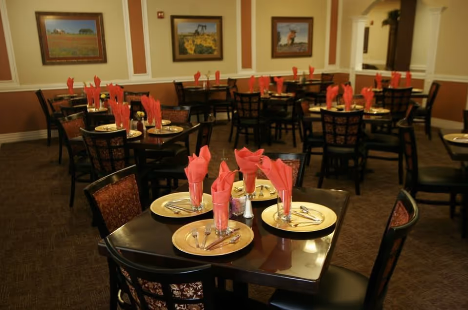 A dining room with multiple dark wood tables set with gold-colored plates, silverware, and glasses holding red folded napkins. The chairs have patterned upholstery, and the walls are decorated with framed landscape paintings. The room has warm lighting and a carpeted floor.