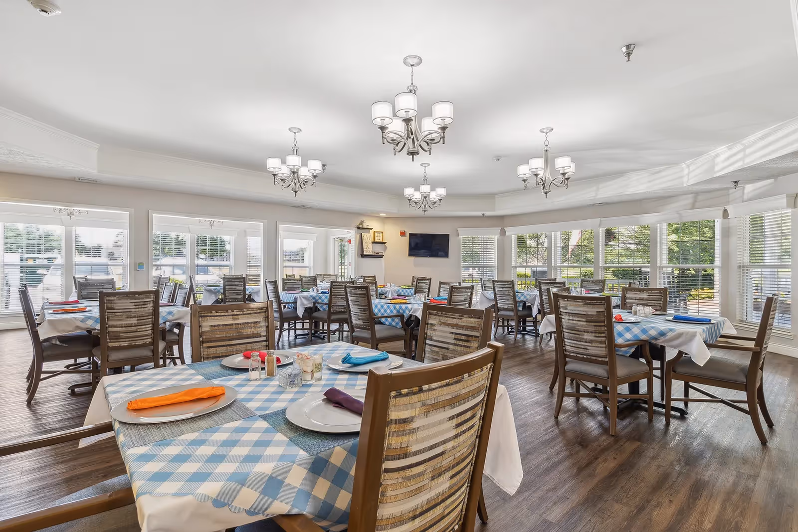 A bright dining room with multiple tables covered in blue and white checkered tablecloths, each set with plates, napkins, and silverware. Wooden chairs surround the tables, and large windows let in natural light, showing greenery outside. Several chandeliers hang from the ceiling, and a flat-screen TV is mounted on one wall.