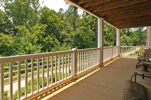 Covered outdoor balcony with white railing overlooking green trees and seating.