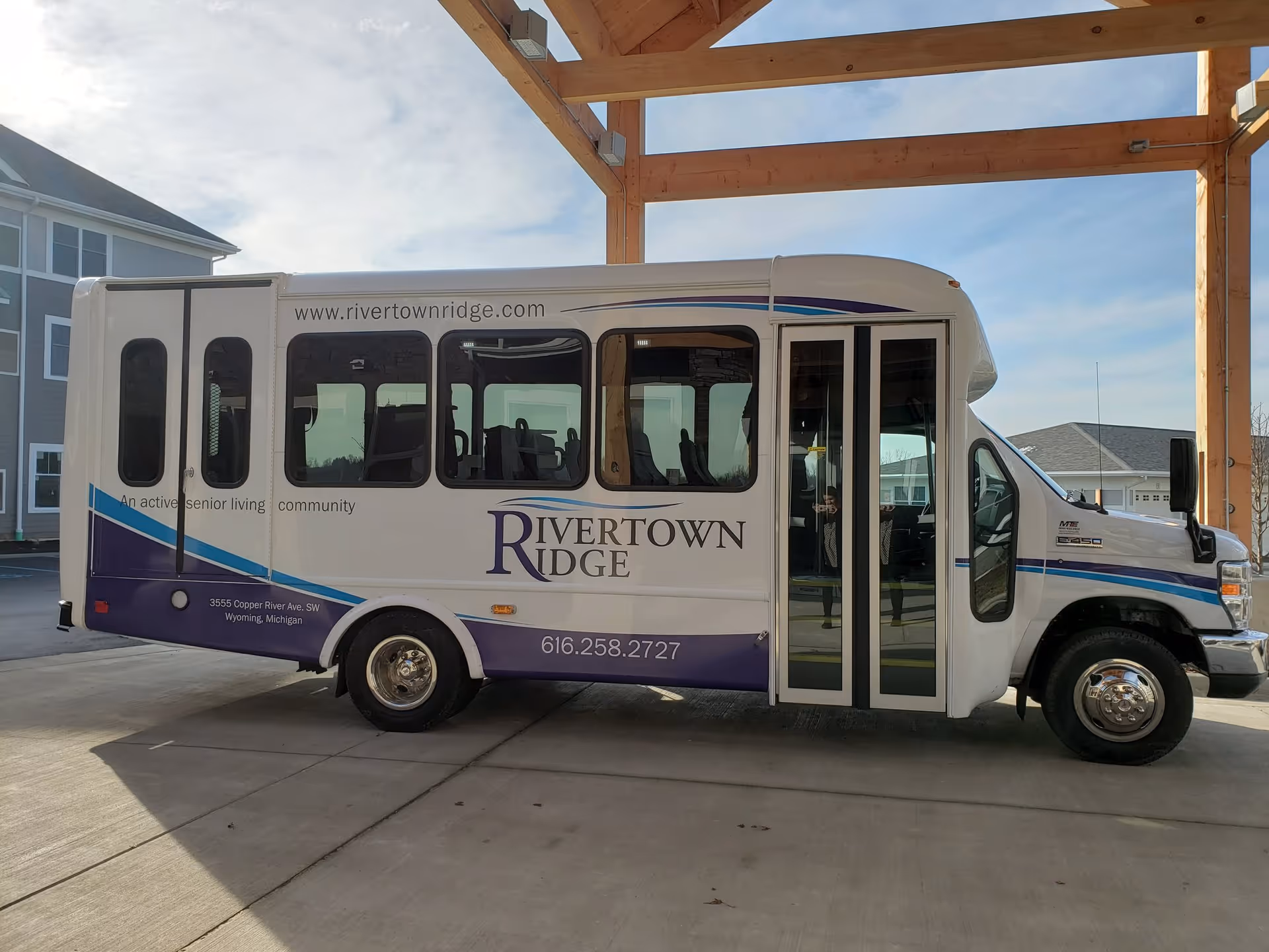 A Rivertown Ridge senior-living shuttle bus parked under a wooden entrance canopy in front of the facility.